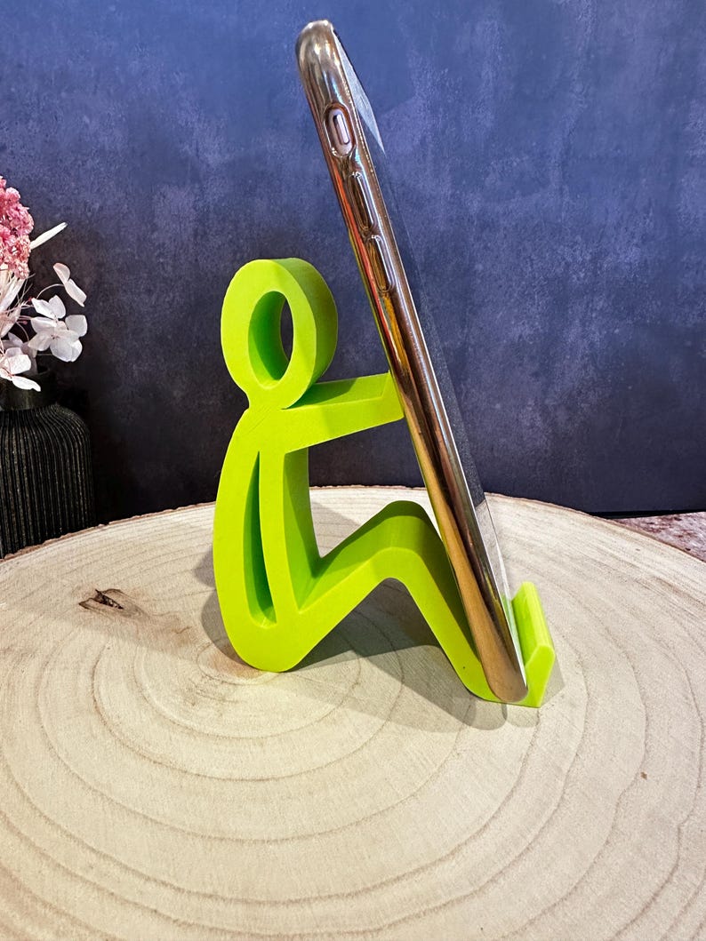 Pu&ograve; includere: Un supporto per telefono verde brillante a forma di persona seduta, che tiene in mano uno smartphone dorato. Il supporto &egrave; realizzato in un materiale solido e si trova su una superficie di legno. Lo sfondo &egrave; blu scuro.