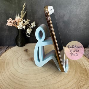 Pu&ograve; includere: Un supporto per telefono azzurro a forma di persona che tiene un telefono con una custodia dorata. Un piccolo vaso nero con fiori secchi &egrave; a sinistra. Il supporto &egrave; su una superficie di legno.