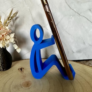 Pu&ograve; includere: Supporto blu per telefono a forma di persona che tiene un telefono dorato. Il supporto &egrave; in plastica e si trova su una superficie di legno. Un vaso nero con fiori secchi sullo sfondo. Altezza: circa 10 cm.