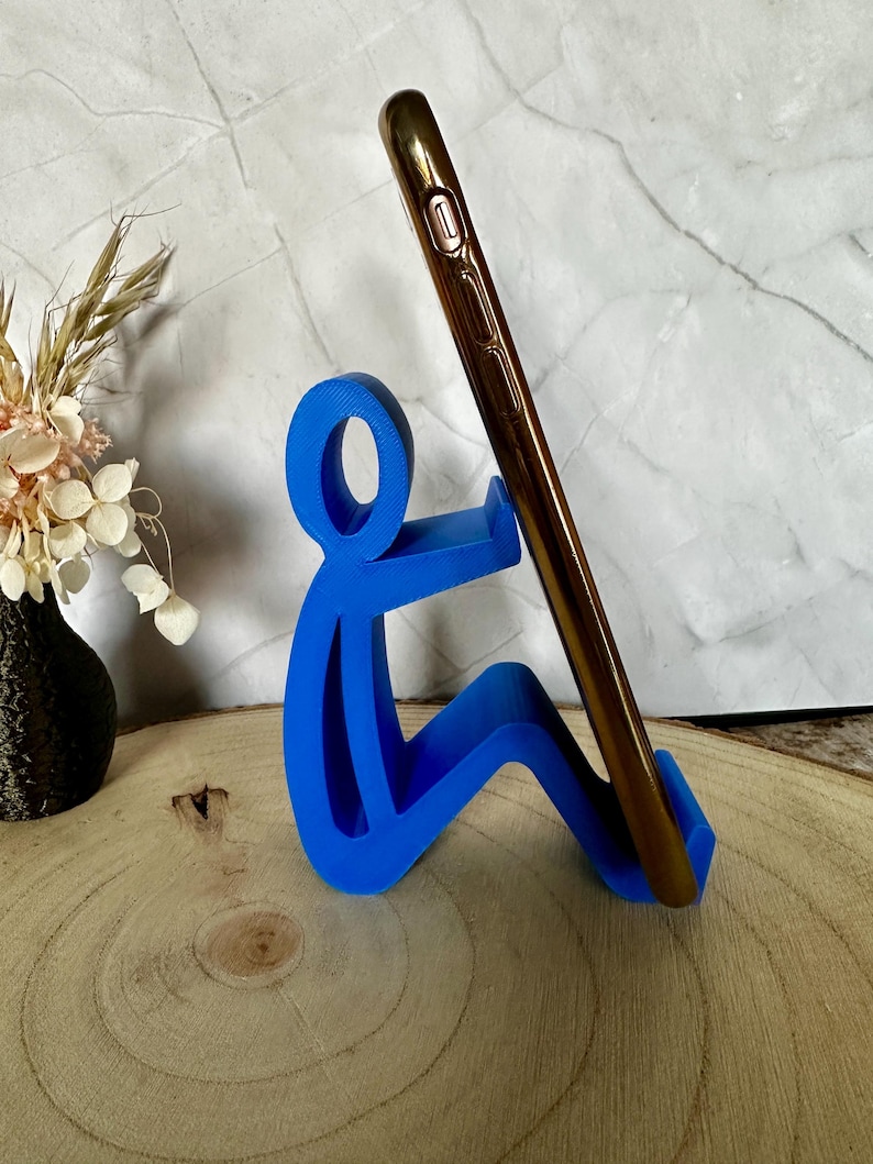 Pu&ograve; includere: Supporto blu per telefono a forma di persona che tiene un telefono dorato. Il supporto &egrave; in plastica e si trova su una superficie di legno. Un vaso nero con fiori secchi sullo sfondo. Altezza: circa 10 cm.
