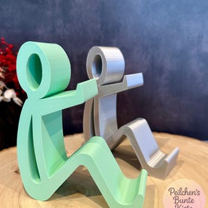 Pu&ograve; includere: Due supporti per telefono astratti a forma di figure sedute. Uno &egrave; verde menta, l'altro argentato. I supporti sono realizzati in un materiale solido e hanno una base curva per sostenere un telefono.