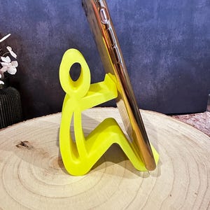 Pu&ograve; includere: Un supporto per telefono giallo neon a forma di persona che tiene in mano uno smartphone dorato. Il supporto &egrave; realizzato in un materiale solido e si trova su una superficie di legno. Il telefono &egrave; angolato per la visualizzazione.