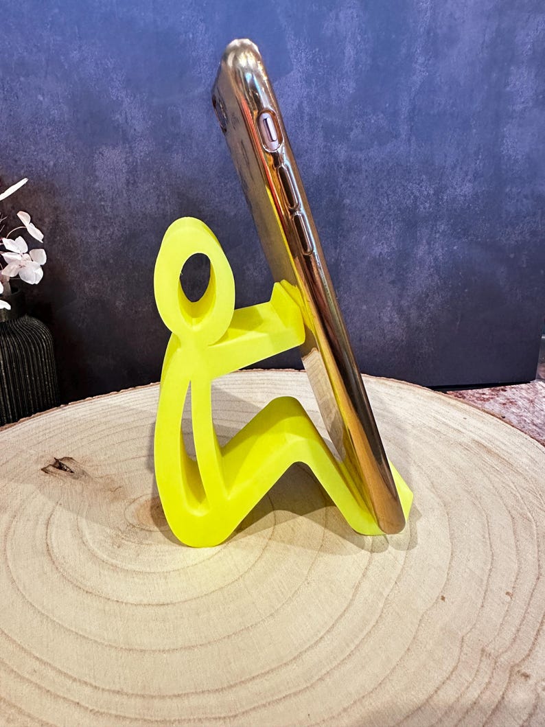 Pu&ograve; includere: Un supporto per telefono giallo neon a forma di persona che tiene in mano uno smartphone dorato. Il supporto &egrave; realizzato in un materiale solido e si trova su una superficie di legno. Il telefono &egrave; angolato per la visualizzazione.