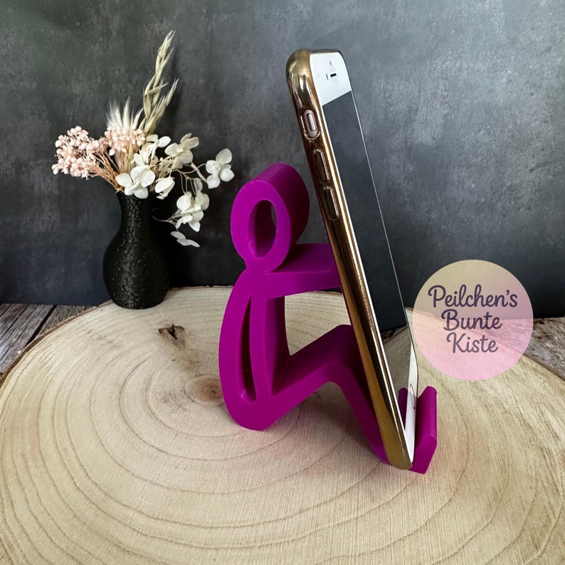Pu&ograve; includere: Un supporto per telefono color magenta a forma di persona seduta, che tiene in mano uno smartphone con cornice dorata. Il supporto &egrave; su una superficie di legno. Un piccolo vaso nero con fiori secchi sullo sfondo.