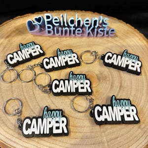 Peut inclure: Six porte-clés noirs avec les mots "happy camper" en lettres blanches avec un accent bleu clair. Les porte-clés sont sur une surface en bois.