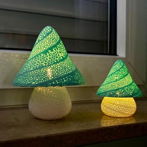 Könnte beinhalten: Zwei dekorative Pilzlampen in Grün und Weiß. Die Lampen haben perforierte, konische Schirme und abgerundete Sockel. Sie sind von innen beleuchtet und erzeugen ein warmes Leuchten. Die Lampen stehen auf einer dunklen Oberfläche in der Nähe eines Fensters.