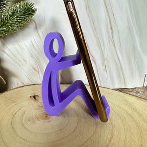 Pu&ograve; includere: Un supporto per telefono viola a forma di persona seduta, che tiene in mano uno smartphone dorato. Il supporto &egrave; realizzato in un materiale solido e si trova su una superficie di legno.