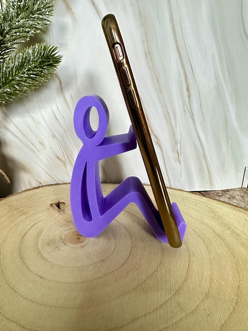 Pu&ograve; includere: Un supporto per telefono viola a forma di persona seduta, che tiene in mano uno smartphone dorato. Il supporto &egrave; realizzato in un materiale solido e si trova su una superficie di legno.