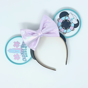 May include: A pair of pink iridescent Minnie Mouse ears with a white bow. The left ear features a blue and white Old Key West Resort logo with a lighthouse and palm trees. The right ear features a blue and white life preserver with a black Mickey Mouse head wearing a sailor hat.
