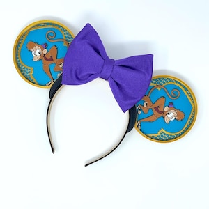 May include: A headband with two round ears featuring a cartoon monkey character on a blue background with gold trim. A large purple bow is centered above the headband. The headband is black.