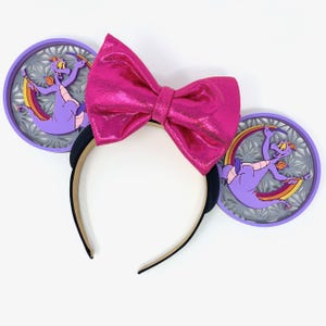May include: Purple and gray Mickey Mouse ears with a pink sparkly bow. The ears feature a purple dragon with a rainbow and a gray snowflake design.
