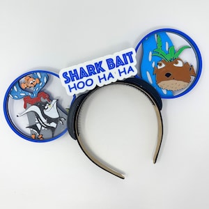 May include: Blue and white Finding Nemo themed Mickey Mouse ears with a black headband. The ears feature a blue and white shark bait sign with the text "Shark Bait Hoo Ha Ha" and a blue and white image of Dory and Nemo.