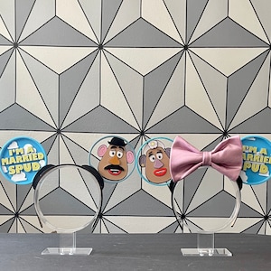 May include: A pair of black Mickey Mouse ears with a blue and white character head on each ear. The left ear has the text "I'm a Married Spud" and the right ear has the text "I'm a Married Pud".