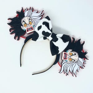 May include: Black headband with Cruella de Vil character ears. The ears feature a black and white cow-print bow and Cruella's face with black and white hair, red accents, and turquoise earrings.