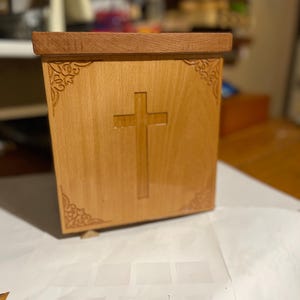 Könnte beinhalten: Eine Holzkiste mit einem Kreuz, das in die Vorderseite geschnitzt ist. Die Kiste besteht aus hellem Holz mit einem dunkleren Holzdeckel. Dekorative Eckverzierungen.