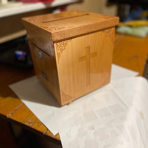 Puede incluir: Una caja de madera de color claro con una ranura en la parte superior y una cruz tallada en la parte delantera. La caja tiene grabados decorativos en las esquinas y una pequeña cerradura de metal. La caja está sobre una hoja de papel blanca.