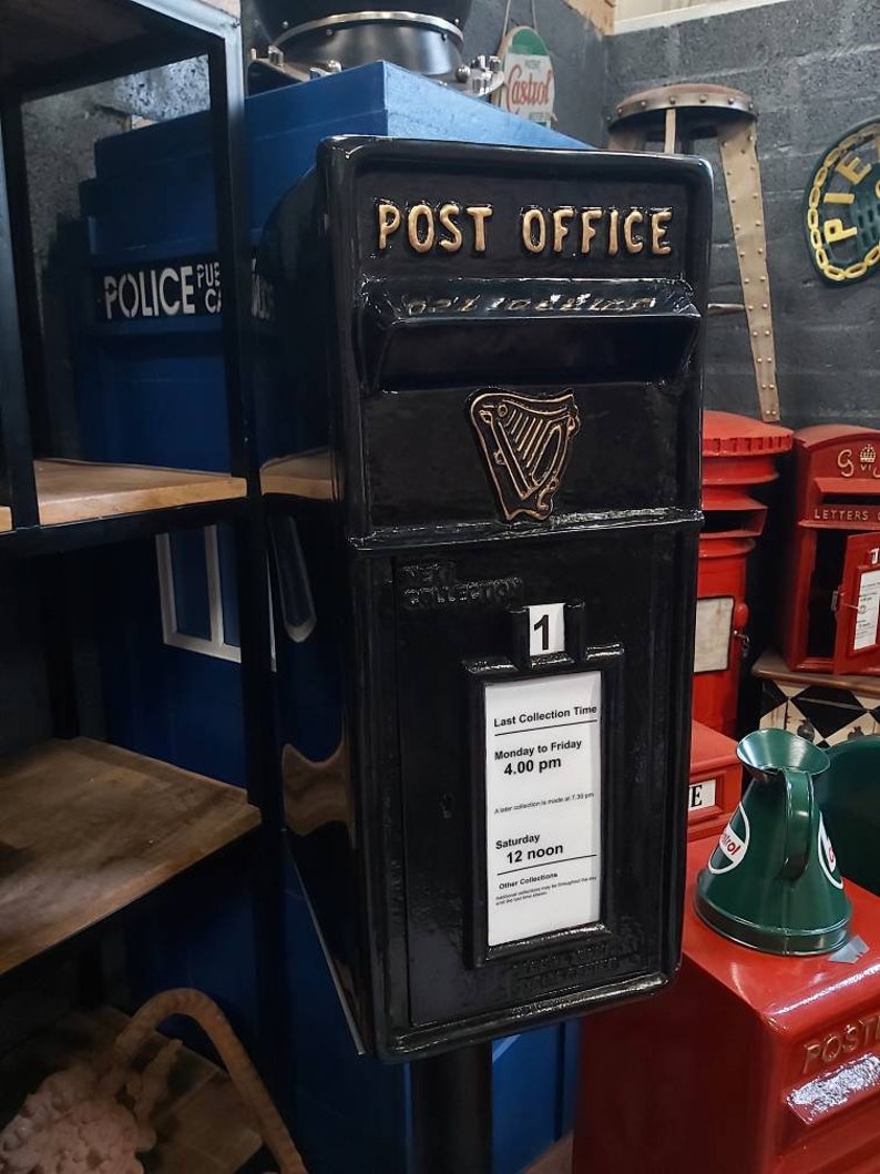 May include: A black cast iron post office mailbox with a gold inscription that reads "POST OFFICE". The mailbox has a gold harp emblem and a white sign with the last collection times. The sign reads "Last Collection Time Monday to Friday 4.00 pm Saturday 12 noon".