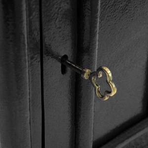 May include: A black metal cabinet door with a keyhole and a gold-colored key inserted. The key has a decorative, flower-shaped head.
