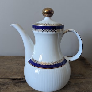 May include: White porcelain coffee pot with a gold and blue trim. The pot has a round body with a spout and a handle. The lid has a gold knob.