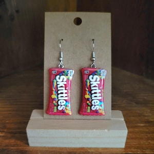 Op de afbeelding: Een paar Skittles snoep oorbellen. Elke oorbel is een miniatuur replica van een Skittles snoepverpakking, met de merknaam en kleurrijke snoep graphics. De oorbellen hangen aan zilverkleurige oorhaakjes.