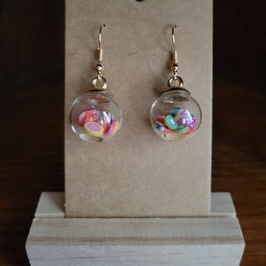 Könnte beinhalten: Ein Paar goldene Ohrhänger mit klaren Glaskugeln, die mit bunten, winzigen Fruchtscheiben gefüllt sind. Die Ohrringe werden auf einem braunen Karton mit Holzsockel präsentiert.