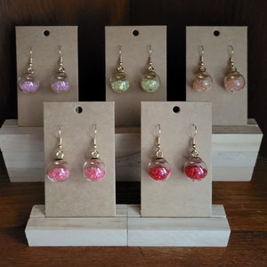 May include: Six pairs of gold dangle earrings with glass teardrop charms filled with colorful glitter. The earrings are displayed on brown card stock.