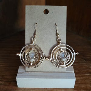 May include: Gold-colored hourglass earrings. Each earring features a circular frame with an hourglass-shaped center filled with sand. The earrings are displayed on a small wooden stand against a neutral background.