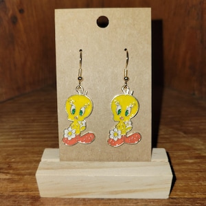 Puede incluir: Un par de pendientes colgantes dorados con el personaje de dibujos animados Piolín. Los pendientes son amarillos con pies naranjas y una flor blanca. Se exhiben en una tarjeta marrón con una base de madera.