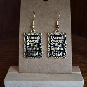 Può includere: Un paio di orecchini pendenti color oro. Ogni orecchino ha un ciondolo rettangolare nero con finiture dorate e il testo "I Solemnly Swear that I'm up to no Good." Gli orecchini sono esposti su una carta marrone.