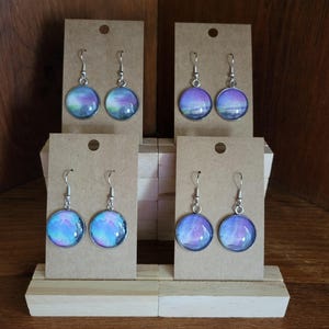 Può includere: Quattro paia di orecchini rotondi color argento con un design astratto e colorato. Ogni orecchino è esposto su un piccolo cartoncino marrone. Gli orecchini presentano un mix di tonalità blu, viola e verde.