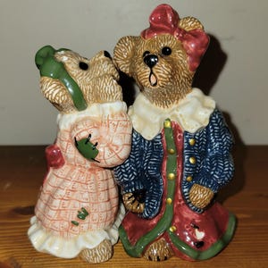 Puede incluir: Un par de saleros y pimenteros de cerámica en forma de osos. Los osos están vestidos con ropa estampada en rojo, verde, azul y blanco. La osa lleva un lazo verde y el oso lleva un lazo rojo.