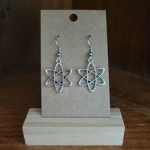 Könnte beinhalten: Ein Paar silberfarbene Ohrringe mit Atom-Design, präsentiert auf einer braunen Karte mit Loch oben, gehalten von einem Holzständer. Der Hintergrund ist in einem warmen, neutralen Ton gehalten. Ein Accessoire mit Wissenschaftsthema.
