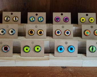 May include: A display of eye-shaped stud earrings in various colors, including yellow, blue, pink, and green. Each pair is mounted on a small, brown cardboard backing. The earrings are arranged on a tiered wooden display.