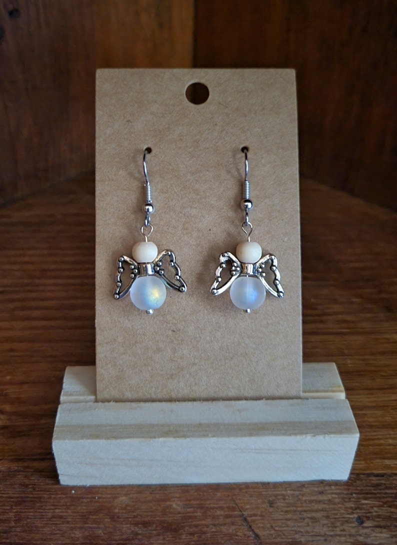 May include: A pair of silver angel-shaped earrings with iridescent white beads and wooden accents.