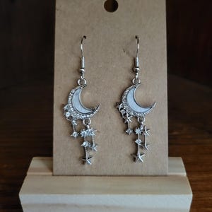 Peut inclure: Boucles d'oreilles en forme de croissant de lune argentées avec des étoiles et des strass pendants. Les boucles d'oreilles sont présentées sur une carte marron.