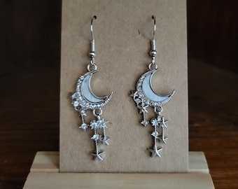 May include: Silver crescent moon earrings with dangling stars and rhinestones. The earrings are displayed on a brown card.