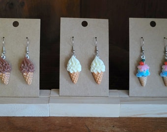 May include: Three pairs of ice cream cone earrings on display. The first pair is brown and white, the second pair is white and orange, and the third pair is pink, blue, and green.