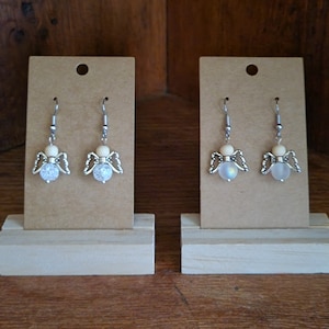 May include: Two pairs of angel-shaped earrings with white beads and silver wings on a brown card display.