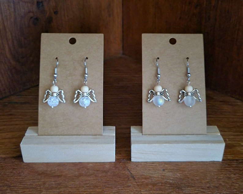 May include: Two pairs of angel-shaped earrings with white beads and silver wings on a brown card display.