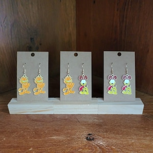 Könnte beinhalten: Drei Paar Ohrhänger mit Zeichentrickfiguren. Zwei Paare zeigen Garfield in Orange, und ein Paar zeigt Odie in Gelb, Rot und Weiß. Die Ohrringe werden auf kleinen, braunen Pappkarten präsentiert.