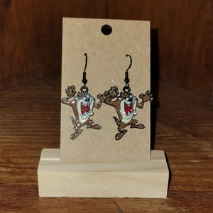 May include: A pair of dangle earrings featuring the Tasmanian Devil cartoon character. The earrings are brown, white, and red with black ear wires. The earrings are displayed on a brown cardboard backing.