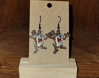 May include: A pair of dangle earrings featuring the Tasmanian Devil cartoon character. The earrings are brown, white, and red with black ear wires. The earrings are displayed on a brown cardboard backing.
