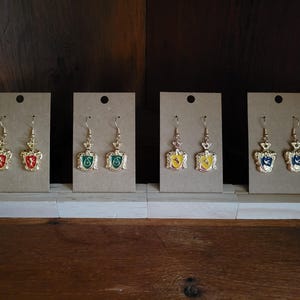 May include: Set of eight gold-tone earrings with enamel charms. Each charm features a different Hogwarts house crest: Gryffindor, Slytherin, Hufflepuff, and Ravenclaw.