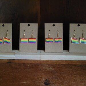 Op de afbeelding: Vier paar regenboogvlag oorbellen weergegeven op individuele kartonnen kaarten. De oorbellen hebben een goudkleurige haak en een rechthoekig vlagontwerp met de kleuren van de regenboog. De oorbellen worden weergegeven op een houten plank.