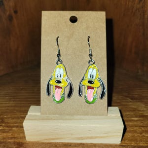 Può includere: Un paio di orecchini pendenti con un design a forma di cane dei cartoni animati. Gli orecchini hanno una faccia gialla, orecchie nere e una lingua rosa. Gli orecchini sono esposti su una carta marrone.