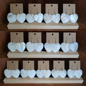Peut inclure: Présentation de boucles d'oreilles en forme de cœur. Les boucles d'oreilles sont blanches avec des reflets irisés et sont suspendues à des crochets argentés. Chaque paire est fixée à une petite carte marron. Les boucles d'oreilles sont disposées sur des étagères en bois.