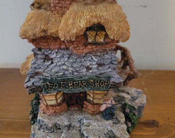 May include: A small, brown, thatched-roof cottage with a brick chimney and a sign that reads "Ted E. Bear Shop". The cottage is made of stone and has a small door and window. There is a small figure of a bear standing in front of the shop.