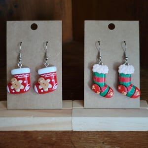 Könnte beinhalten: Zwei Paar Weihnachtsstrumpf-Ohrringe. Ein Paar ist rot mit einem Lebkuchenmann-Design und das andere Paar ist grün-rot gestreift mit weißem Fellbesatz.