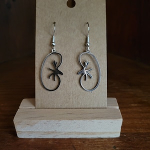 Op de afbeelding: Een paar zilveren nierenvormige oorbellen met een gedetailleerd uitgesneden ontwerp. De oorbellen worden gepresenteerd op een bruine kaart met een houten basis.