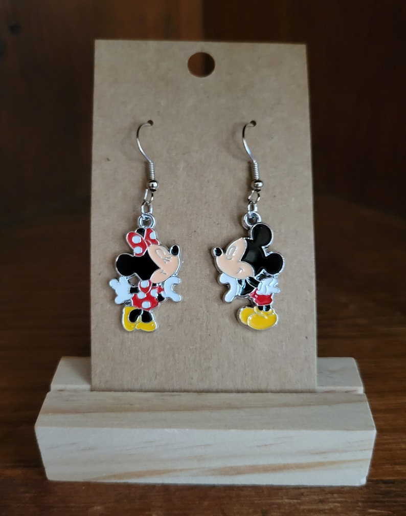 K&ouml;nnte beinhalten: Ein Paar Ohrh&auml;nger mit Mickey und Minnie Mouse. Die Ohrringe haben silberfarbene Haken und sind aus schwarzem, rotem, wei&szlig;em und gelbem Emaille gefertigt. Die Ohrringe werden auf einer braunen Karte pr&auml;sentiert.